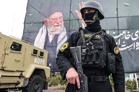 Armed Iranian security officer standing in front of an armored vehicle with a large mural of a national leader in the background during wartime